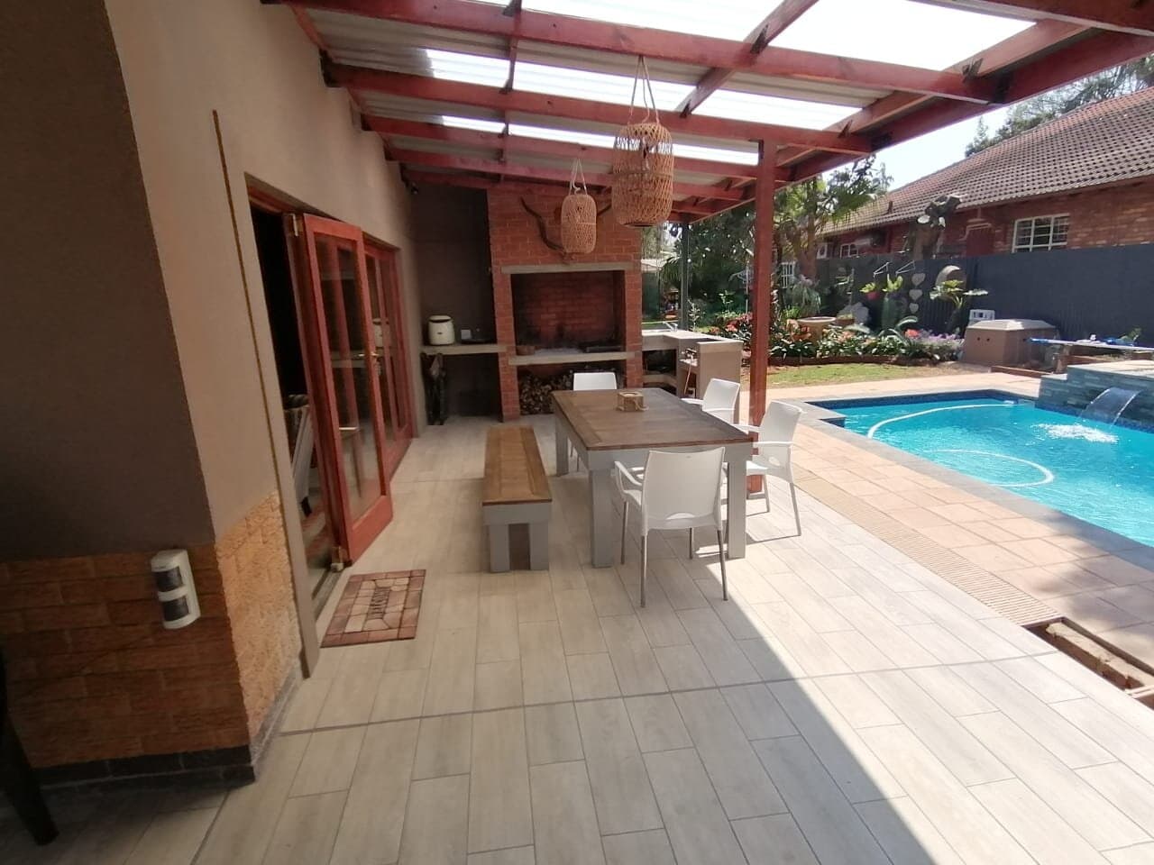 Covered timber patio with a built-in braai, dining table and pool with water feature
